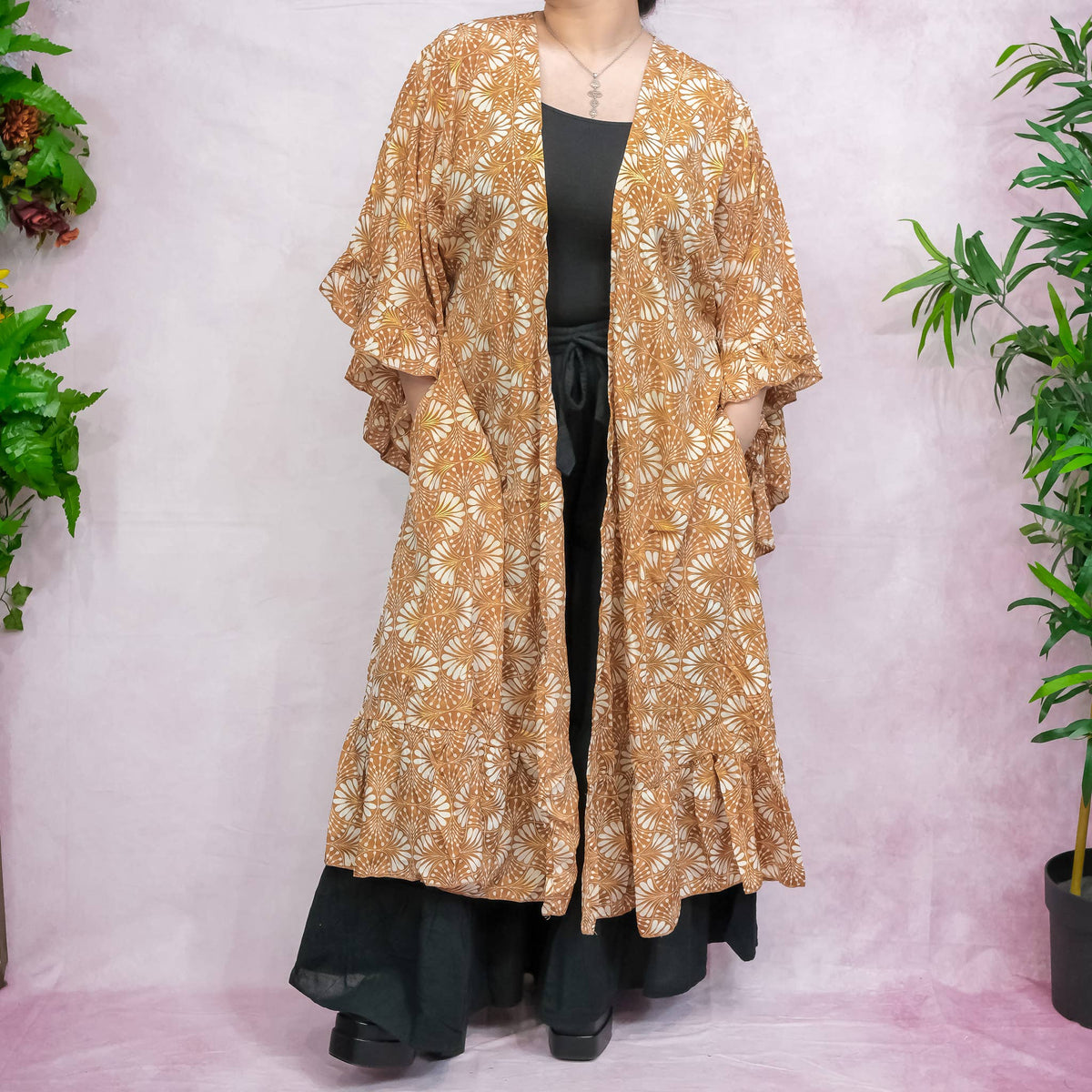 Long beige floral patterned dress over black pants on a white background with plants.