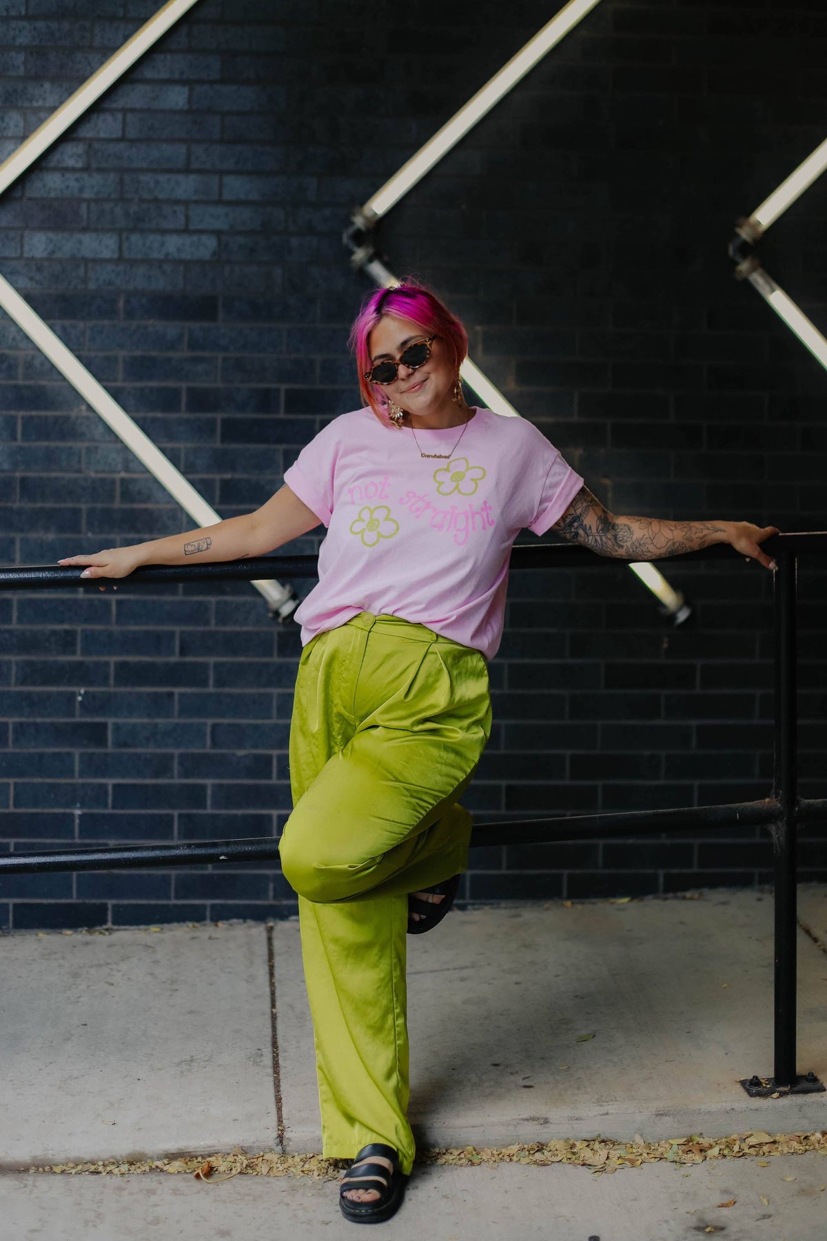 Person wearing a pink t-shirt and green pants leaning against a black metal railing with a dark tiled wall background.