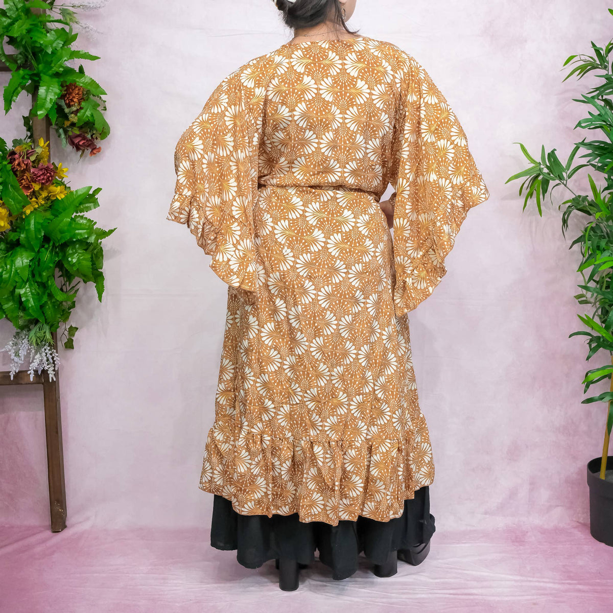 Person wearing a gold floral dress with a white background and plants on either side.