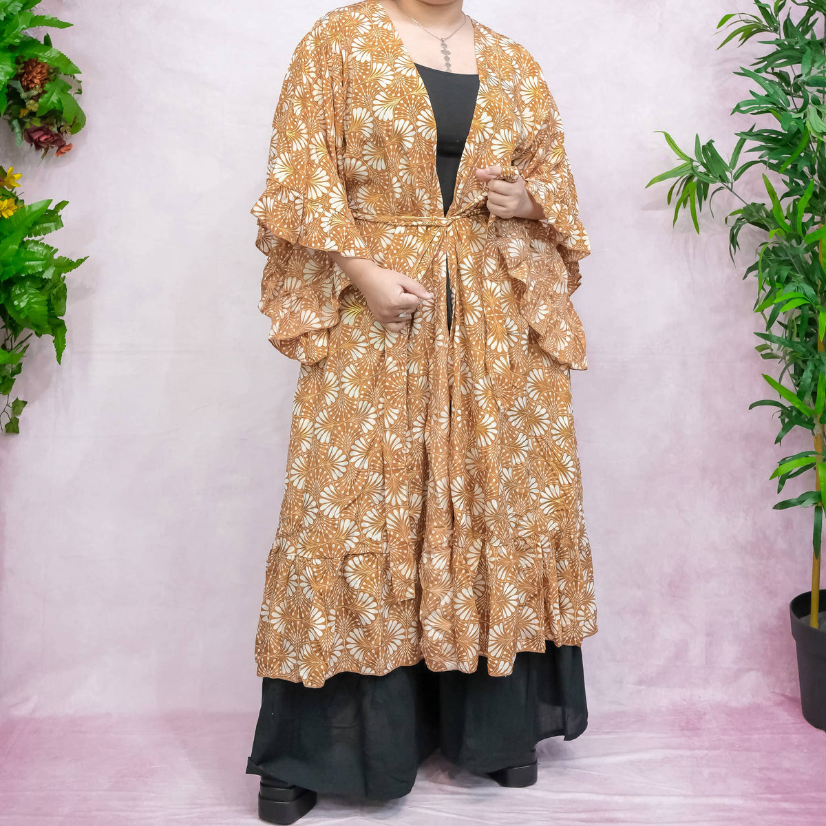 Person wearing a long, patterned beige coat over a black outfit against a white background with plants.