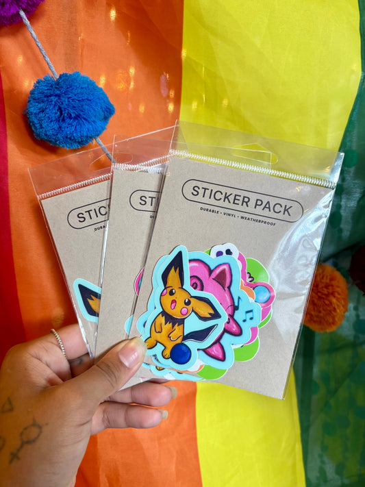 A hand holds three packaged sticker packs in front of a bright rainbow-colored fabric backdrop. The front pack shows a stack of colorful cartoon-style stickers, including a yellow creature and pink and blue characters, on a kraft paper backing card labeled 'STICKER PACK'.