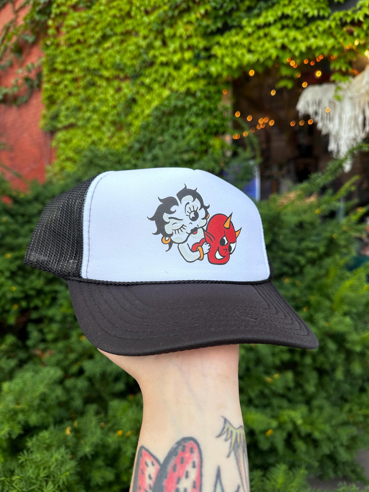 A hand holding a black and white trucker hat with a graphic design of Betty Boop and the Hot Stuff Devil on it.
