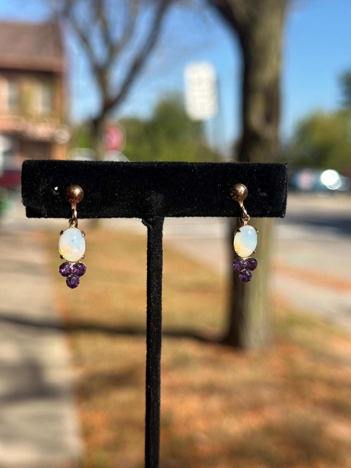 Pair of earrings with opal and purple gemstones on a black stand against a blurred outdoor background.