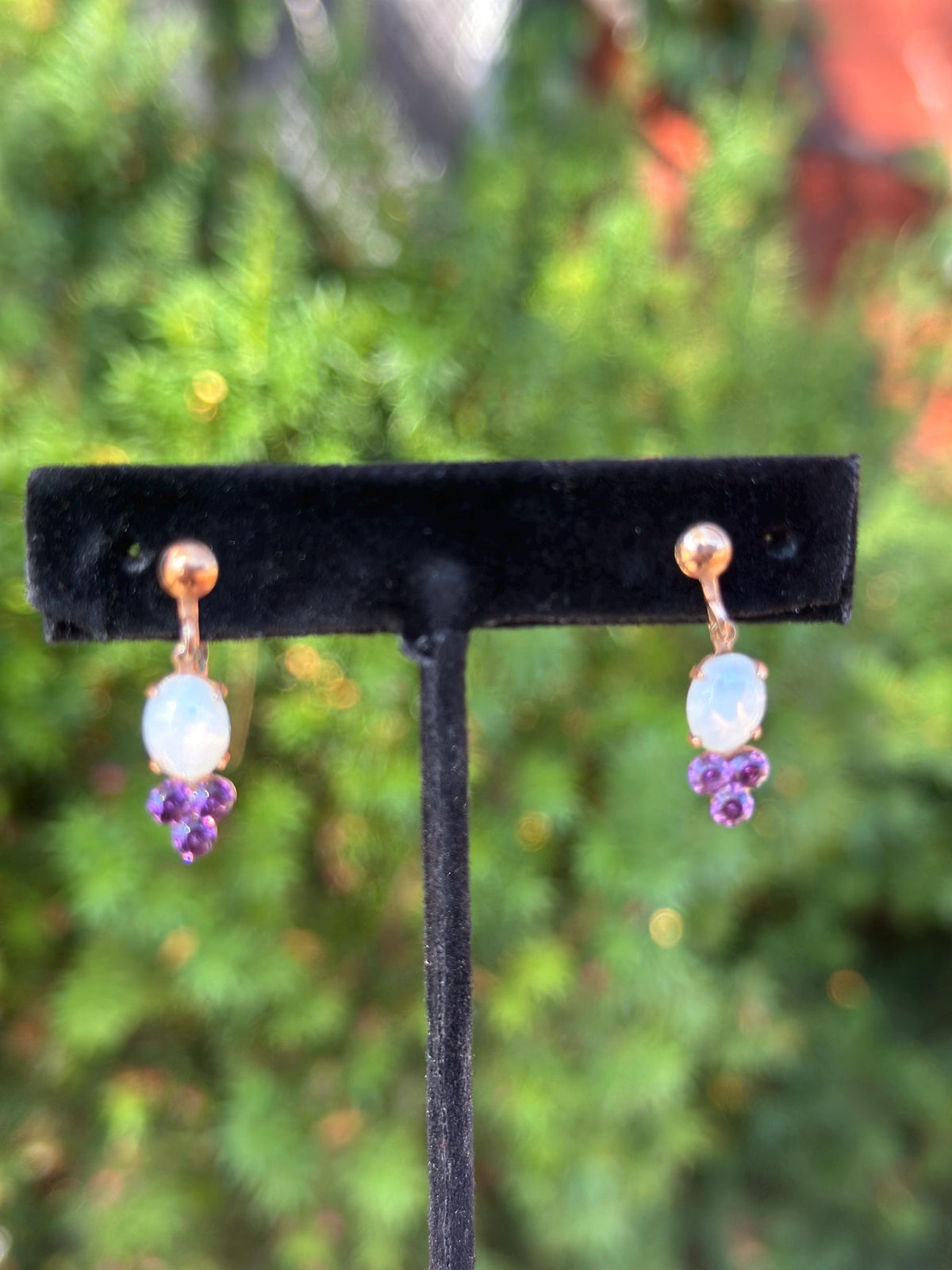 Pair of earrings with opal and purple gemstones on a black stand against a blurred green background