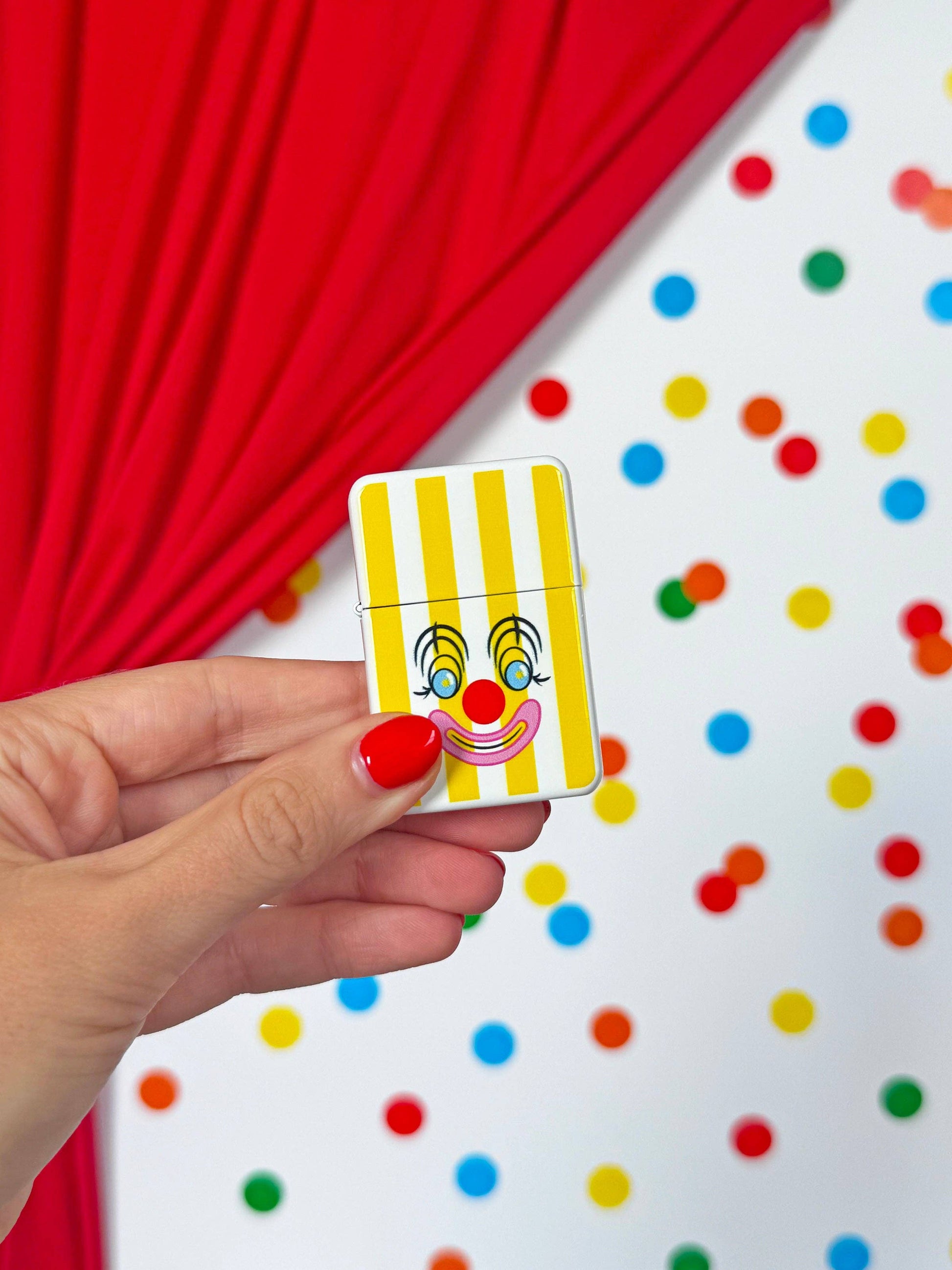 Hand holding a small rectangular object with a clown face design against a colorful polka dot background.