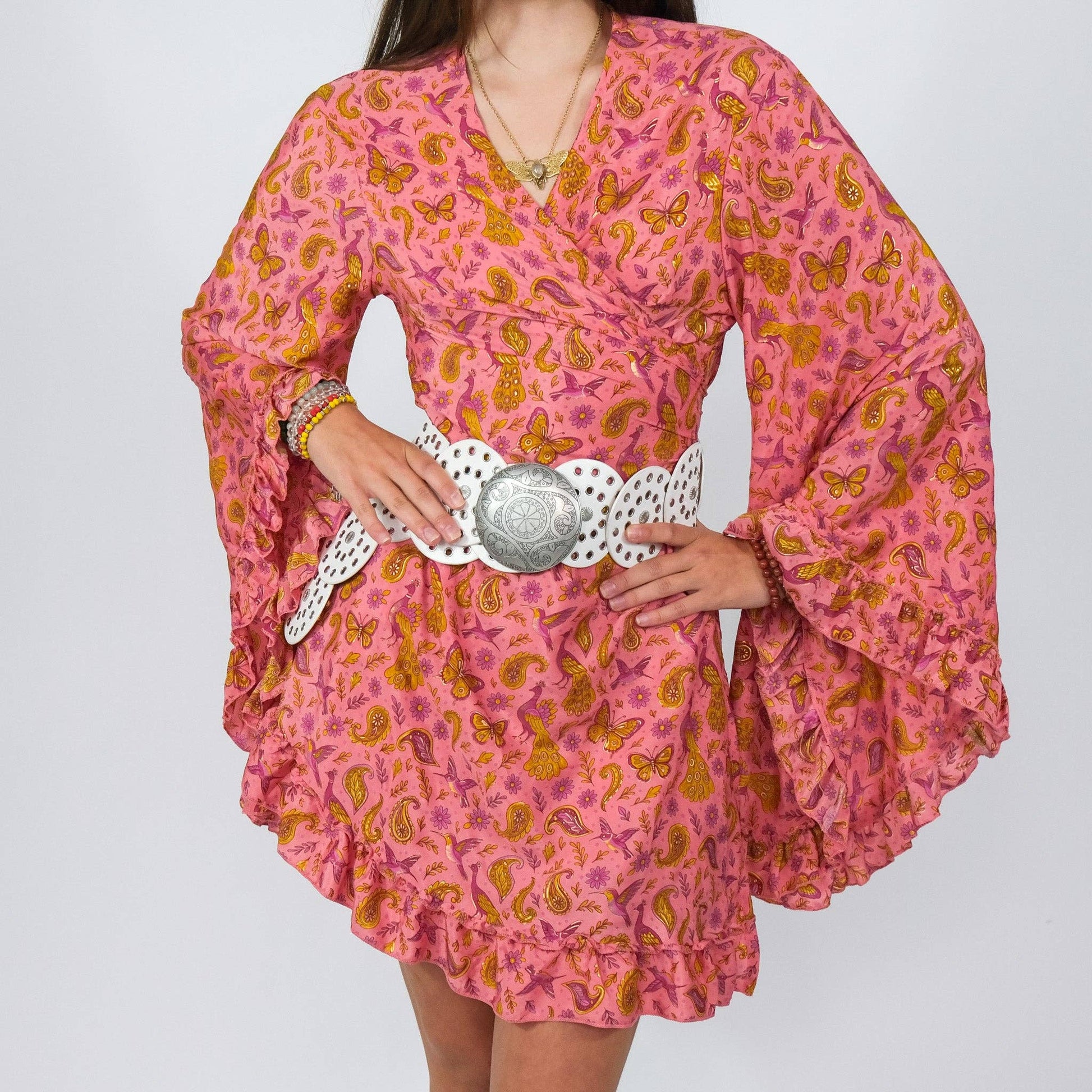 Person wearing a pink dress with orange patterns and a decorative belt on a plain background