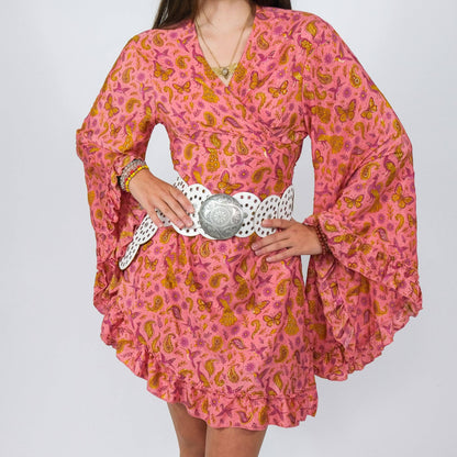 Person wearing a pink dress with orange patterns and a decorative belt on a plain background