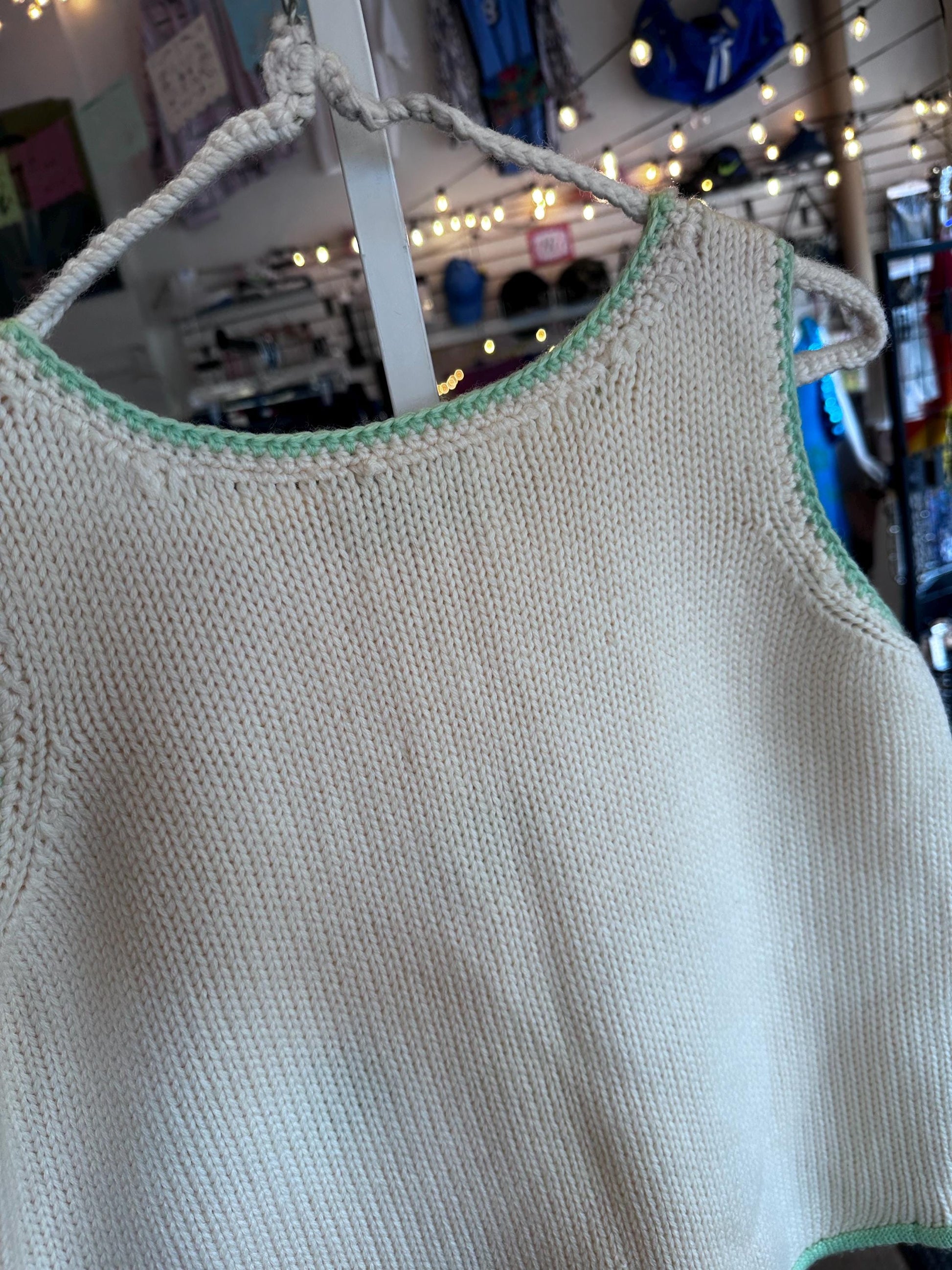 Light-colored sleeveless top with green trim on a hanger against a blurred store background.