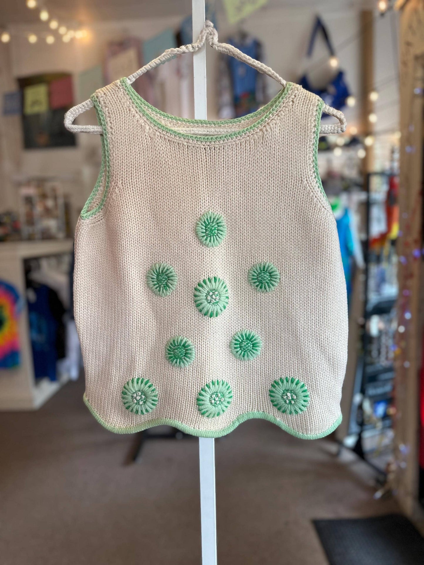Beige knitted tank top with green floral patterns on a hanger in a store setting.