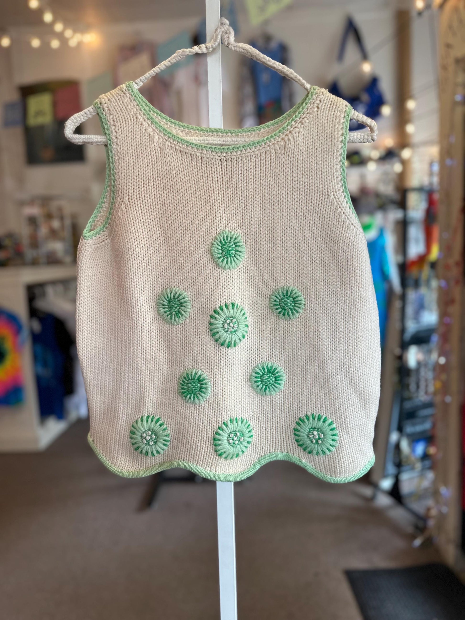 Beige sleeveless top with green floral patterns on a hanger against a blurred indoor background.