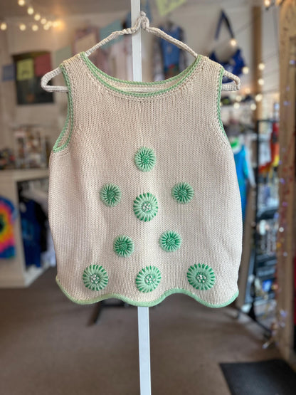Beige sleeveless top with green floral patterns on a hanger against a blurred indoor background.