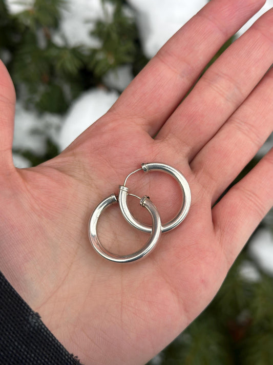 Two silver hoop earrings held in a hand with a blurred natural background